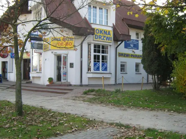 Budynek z żółtymi banerami "OKNA DRZWI" i "DRZWI PARAPETY", z drzewem po lewej.